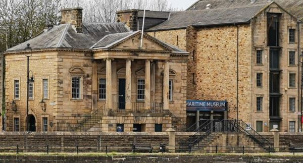 Photo of Lancaster Maritime Museum, which consists of two connected buildings: the grand Georgian custom house with a columned portico on the left and a tall warehouse on the right. Both are built from golden sandstone.